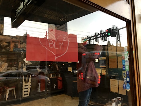 A reflection on a glass window shows a person inside a building next to a red poster with 'BLM' written inside a heart. The background reveals city infrastructure, including a road with cars and traffic lights.