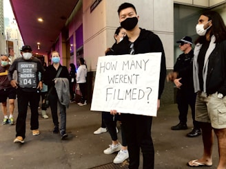 A group of people, some wearing masks, gather on a city street. One individual at the center holds a sign reading 'How many weren't filmed?' indicating a protest or demonstration. A police officer is visible in the background.