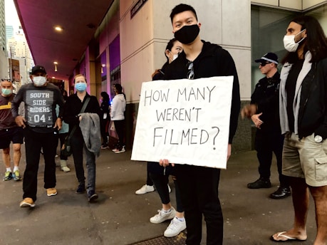 A group of people, some wearing masks, gather on a city street. One individual at the center holds a sign reading 'How many weren't filmed?' indicating a protest or demonstration. A police officer is visible in the background.