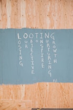 A painted section on a wooden board featuring vertically arranged words forming an acronym: LOOTING, with each letter starting a word. The words are written in white paint on a sky-blue background, contrasting with the natural wood color around it.