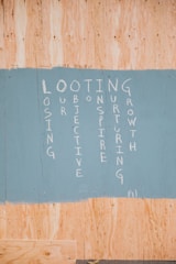 A painted section on a wooden board featuring vertically arranged words forming an acronym: LOOTING, with each letter starting a word. The words are written in white paint on a sky-blue background, contrasting with the natural wood color around it.