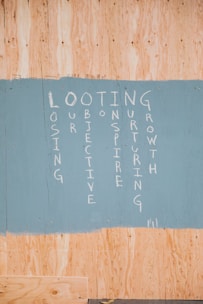 A painted section on a wooden board featuring vertically arranged words forming an acronym: LOOTING, with each letter starting a word. The words are written in white paint on a sky-blue background, contrasting with the natural wood color around it.