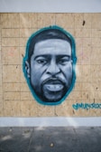 A mural painted on wooden panels features a grayscale portrait of a man's face highlighted with a teal outline. Peace symbols are drawn faintly in pink on each side. The artwork includes a signature with a heart in teal color in the bottom right corner.