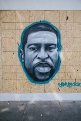A mural painted on wooden panels features a grayscale portrait of a man's face highlighted with a teal outline. Peace symbols are drawn faintly in pink on each side. The artwork includes a signature with a heart in teal color in the bottom right corner.