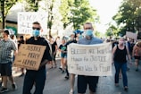 A group of people are participating in a protest march. They are holding signs with messages advocating for justice and equality. The scene is set on a sunny day in an urban area, with trees lining the street, and the participants are wearing face masks. The crowd appears diverse and focused on their cause.