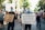A group of people are participating in a protest march. They are holding signs with messages advocating for justice and equality. The scene is set on a sunny day in an urban area, with trees lining the street, and the participants are wearing face masks. The crowd appears diverse and focused on their cause.