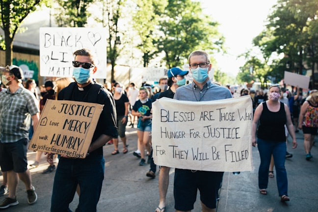 A group of people are participating in a protest march. They are holding signs with messages advocating for justice and equality. The scene is set on a sunny day in an urban area, with trees lining the street, and the participants are wearing face masks. The crowd appears diverse and focused on their cause.