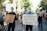 A group of people are participating in a protest march. They are holding signs with messages advocating for justice and equality. The scene is set on a sunny day in an urban area, with trees lining the street, and the participants are wearing face masks. The crowd appears diverse and focused on their cause.