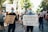 A group of people are participating in a protest march. They are holding signs with messages advocating for justice and equality. The scene is set on a sunny day in an urban area, with trees lining the street, and the participants are wearing face masks. The crowd appears diverse and focused on their cause.