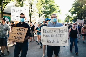 A group of people are participating in a protest march. They are holding signs with messages advocating for justice and equality. The scene is set on a sunny day in an urban area, with trees lining the street, and the participants are wearing face masks. The crowd appears diverse and focused on their cause.