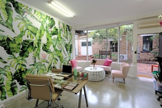 A modern office or workspace featuring a vibrant green leaf-patterned wallpaper on one wall, adding a tropical aesthetic to the room. A glass desk is equipped with a laptop, a notebook, and several desk accessories. In front of a large window, which allows natural light, there is a round white coffee table surrounded by pink cushioned chairs. The window overlooks a courtyard with brickwork and some greenery. The space is decorated with elements like indoor plants and a ceiling-mounted light, creating a fresh and inviting atmosphere.
