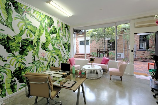 A modern office or workspace featuring a vibrant green leaf-patterned wallpaper on one wall, adding a tropical aesthetic to the room. A glass desk is equipped with a laptop, a notebook, and several desk accessories. In front of a large window, which allows natural light, there is a round white coffee table surrounded by pink cushioned chairs. The window overlooks a courtyard with brickwork and some greenery. The space is decorated with elements like indoor plants and a ceiling-mounted light, creating a fresh and inviting atmosphere.