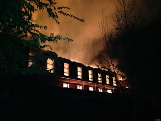 A large building is engulfed in flames during nighttime, with bright orange and yellow fire illuminating multiple windows and thick smoke billowing into the dark sky. Shadows of surrounding trees are faintly visible against the intense glow of the fire.