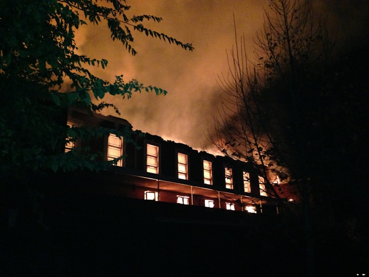 A large building is engulfed in flames during nighttime, with bright orange and yellow fire illuminating multiple windows and thick smoke billowing into the dark sky. Shadows of surrounding trees are faintly visible against the intense glow of the fire.