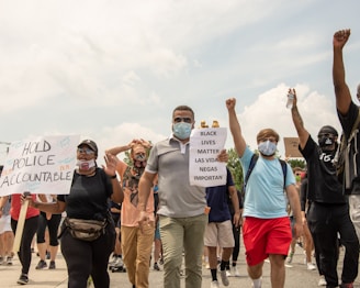 A diverse group of people are walking together, holding signs with messages such as 'HOLD POLICE ACCOUNTABLE' and 'BLACK LIVES MATTER LAS VIDAS NEGRAS IMPORTAN.' The group includes individuals of different genders and ethnicities wearing masks, with some raising their fists in solidarity. The sky in the background is cloudy.