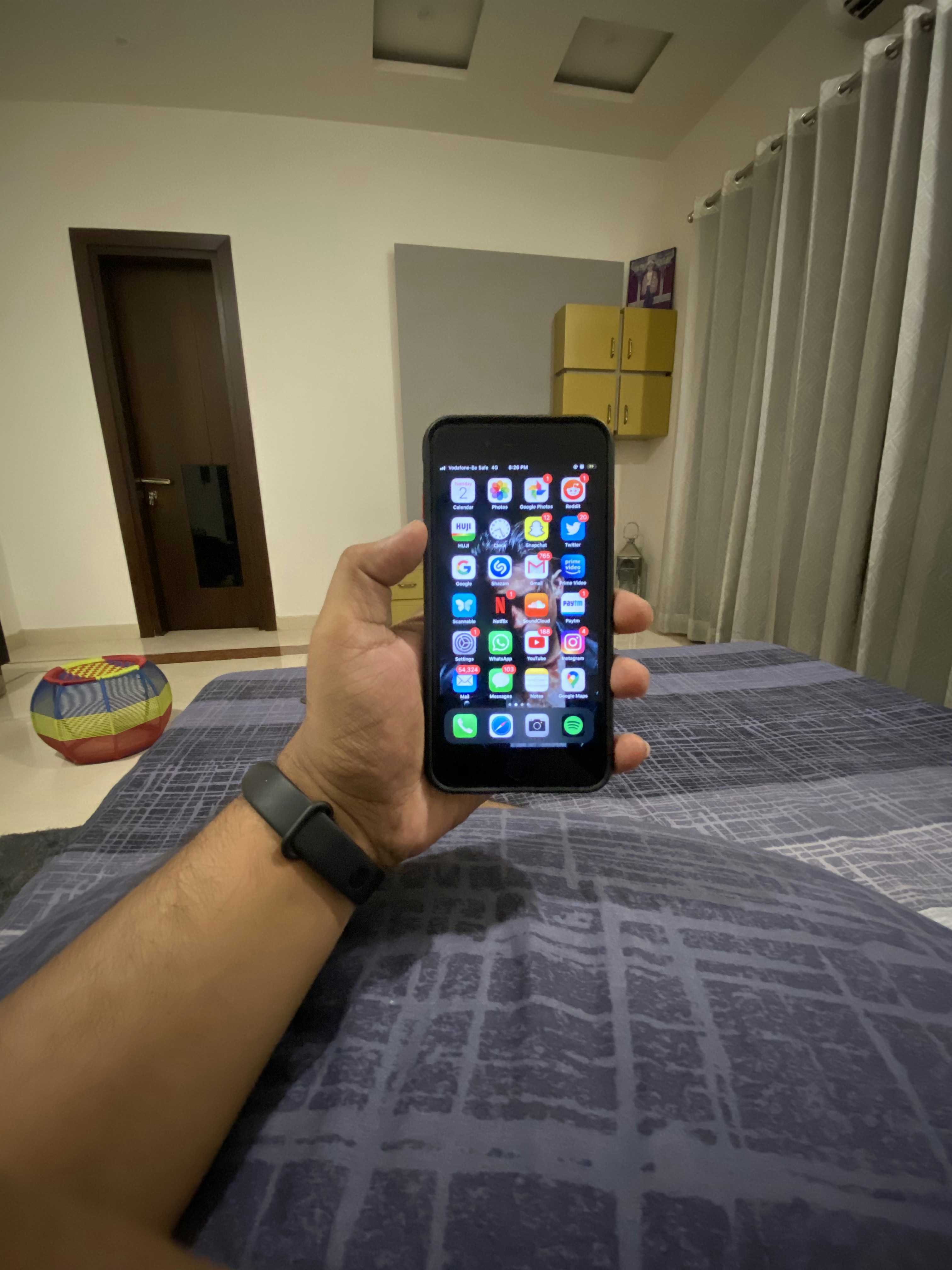 A hand holding a smartphone displaying a grid of colorful application icons. The room features modern decor with a grey curtain, a door, and a cushioned stool with a multi-colored cover. A smartwatch is worn on the wrist holding the phone, and a soft light illuminates the space.