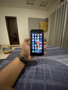 A hand holding a smartphone displaying a grid of colorful application icons. The room features modern decor with a grey curtain, a door, and a cushioned stool with a multi-colored cover. A smartwatch is worn on the wrist holding the phone, and a soft light illuminates the space.