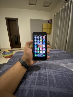 A hand holding a smartphone displaying a grid of colorful application icons. The room features modern decor with a grey curtain, a door, and a cushioned stool with a multi-colored cover. A smartwatch is worn on the wrist holding the phone, and a soft light illuminates the space.