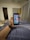 A hand holding a smartphone displaying a grid of colorful application icons. The room features modern decor with a grey curtain, a door, and a cushioned stool with a multi-colored cover. A smartwatch is worn on the wrist holding the phone, and a soft light illuminates the space.