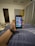 A hand holding a smartphone displaying a grid of colorful application icons. The room features modern decor with a grey curtain, a door, and a cushioned stool with a multi-colored cover. A smartwatch is worn on the wrist holding the phone, and a soft light illuminates the space.