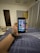 A hand holding a smartphone displaying a grid of colorful application icons. The room features modern decor with a grey curtain, a door, and a cushioned stool with a multi-colored cover. A smartwatch is worn on the wrist holding the phone, and a soft light illuminates the space.