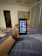 A hand holding a smartphone displaying a grid of colorful application icons. The room features modern decor with a grey curtain, a door, and a cushioned stool with a multi-colored cover. A smartwatch is worn on the wrist holding the phone, and a soft light illuminates the space.