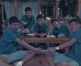 Five young men wearing similar outfits are sitting around a wooden table in a living room. They express playful and humorous facial expressions, with one pretending to pull a remote control from another's hand. The room is decorated with curtains, various items on the table, and household objects in the background.