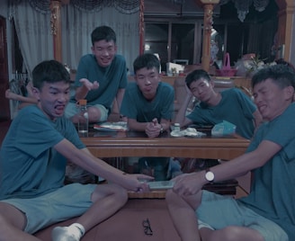 Five young men wearing similar outfits are sitting around a wooden table in a living room. They express playful and humorous facial expressions, with one pretending to pull a remote control from another's hand. The room is decorated with curtains, various items on the table, and household objects in the background.