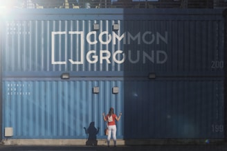 A woman with long hair wearing a red top and white pants stands in front of a large blue shipping container. The container has the words 'Common Ground' painted on it in large white letters. There are also smaller white letters that say 'Retail / F&B Activity.' The scene is partly in shadow with a distinct line of sunlight separating the light and dark areas.
