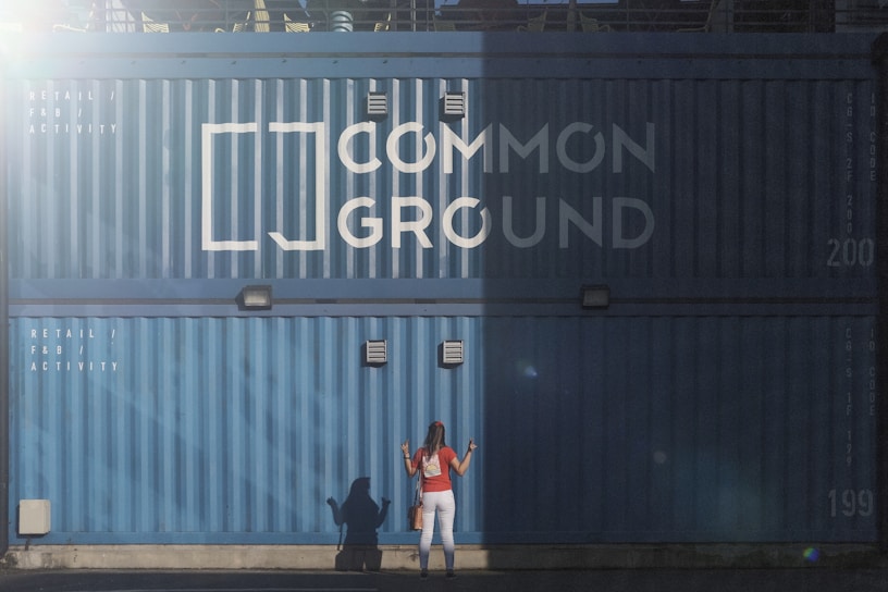 A woman with long hair wearing a red top and white pants stands in front of a large blue shipping container. The container has the words 'Common Ground' painted on it in large white letters. There are also smaller white letters that say 'Retail / F&B Activity.' The scene is partly in shadow with a distinct line of sunlight separating the light and dark areas.