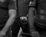 Three individuals in protective gear are partially visible, with one in the center holding a small canister. Their attire suggests a law enforcement or security role, and the image has a serious mood.