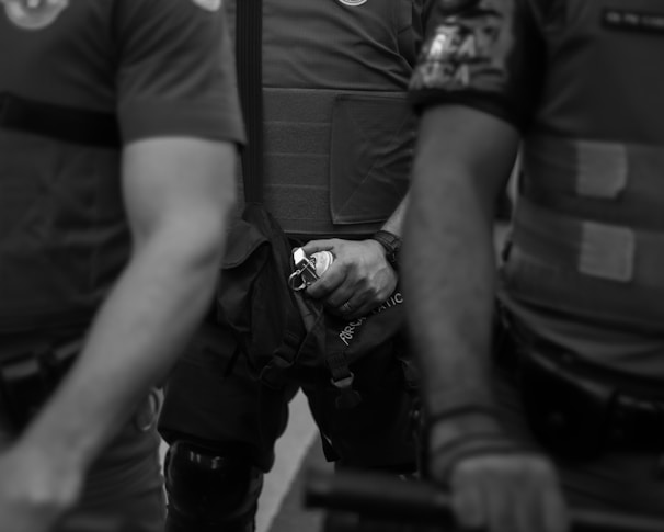 Three individuals in protective gear are partially visible, with one in the center holding a small canister. Their attire suggests a law enforcement or security role, and the image has a serious mood.