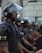 A group of uniformed individuals wearing protective helmets and face shields stands in a line. Each wears dark gear with visible yellow lettering on the sleeves, suggesting a force or security group. The scene appears serious and organized, taking place in a city environment.