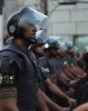 A group of uniformed individuals wearing protective helmets and face shields stands in a line. Each wears dark gear with visible yellow lettering on the sleeves, suggesting a force or security group. The scene appears serious and organized, taking place in a city environment.