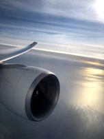 An airplane engine is seen in close-up with one of the wings extending gracefully into the horizon. The background is composed of a soft, cloudy sky with the sun's reflection creating a golden shimmer on the water below.