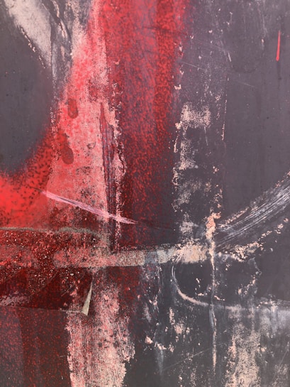 An abstract composition featuring dynamic strokes and splashes of red and black paint. The surface is textured with areas of light beige and hints of white, creating a dramatic contrast. The image conveys a sense of movement and intensity through its layered application of color.