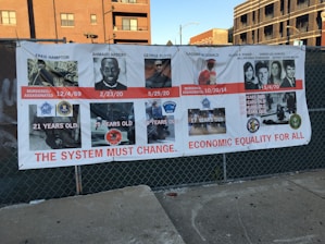 A large banner with images of individuals, alongside details such as their names, dates of death, ages, and associated incidents. It displays messages advocating for systemic change and economic equality. The banner is attached to a fence, with buildings visible in the background.