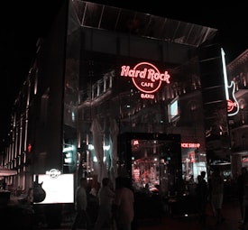 The image captures a Hard Rock Cafe at night, with a prominent red neon sign of the establishment reflecting off the glass building. Pedestrians move along the sidewalk in front, and the glow from various other illuminated signs and lights creates an urban nightlife ambiance.