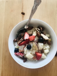 A bowl of oatmeal topped with an assortment of fresh fruits and nuts. The toppings include strawberry slices, blueberries, apple pieces, almonds, walnuts, and shredded coconut. A spoon is placed in the bowl, which is situated on a wooden table.