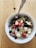 A bowl of oatmeal topped with an assortment of fresh fruits and nuts. The toppings include strawberry slices, blueberries, apple pieces, almonds, walnuts, and shredded coconut. A spoon is placed in the bowl, which is situated on a wooden table.