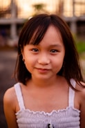 A young girl with long dark hair and a gentle smile, wearing a white sleeveless top. The background is softly blurred with hints of greenery and structures.