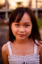 A young girl with long dark hair and a gentle smile, wearing a white sleeveless top. The background is softly blurred with hints of greenery and structures.