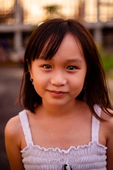 A young girl with long dark hair and a gentle smile, wearing a white sleeveless top. The background is softly blurred with hints of greenery and structures.