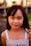 A young girl with long dark hair and a gentle smile, wearing a white sleeveless top. The background is softly blurred with hints of greenery and structures.