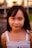 A young girl with long dark hair and a gentle smile, wearing a white sleeveless top. The background is softly blurred with hints of greenery and structures.