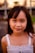 A young girl with long dark hair and a gentle smile, wearing a white sleeveless top. The background is softly blurred with hints of greenery and structures.