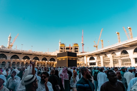 A large crowd of people, many dressed in white, are gathered around the Kaaba, a cuboid building central to the Islamic faith, located in the courtyard of the Great Mosque of Mecca. High cranes and scaffolding suggest ongoing construction work surrounding the mosque. The sky is clear and blue, adding to the bright daylight illuminating the scene.