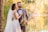 A bride and groom are standing together in a scenic outdoor setting by a lake. The bride is wearing a white lace wedding dress with a long veil, and the groom is in a gray suit with a bow tie. The bride holds a colorful bouquet of flowers. The couple is surrounded by lush greenery and trees displaying autumn colors, with a calm body of water in the background reflecting the foliage.
