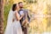 A bride and groom are standing together in a scenic outdoor setting by a lake. The bride is wearing a white lace wedding dress with a long veil, and the groom is in a gray suit with a bow tie. The bride holds a colorful bouquet of flowers. The couple is surrounded by lush greenery and trees displaying autumn colors, with a calm body of water in the background reflecting the foliage.