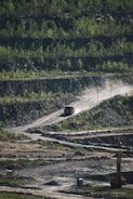 A large truck drives along a winding dirt road surrounded by green vegetation and terraced rocky hillsides. The setting appears to be an open-pit mine or quarry, with large areas of exposed rock and sparse vegetation.